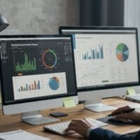 Persoon werkt aan een bureau met drie monitoren waarop grafieken, diagrammen en bedrijfsrapporten worden weergegeven; afgedrukte diagrammen en plakbriefjes zijn zichtbaar in de werkruimte.