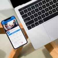 Een smartphone met een Facebook-pagina ligt naast het toetsenbord van een laptop op een houten bureau.