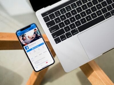 Een smartphone met een Facebook-pagina ligt naast het toetsenbord van een laptop op een houten bureau.