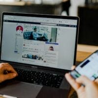 Persoon met een laptop die een Facebook-profielpagina weergeeft terwijl hij een smartphone vasthoudt, beide op een houten tafel.