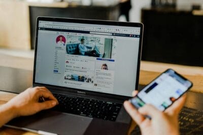 Persoon met een laptop die een Facebook-profielpagina weergeeft terwijl hij een smartphone vasthoudt, beide op een houten tafel.