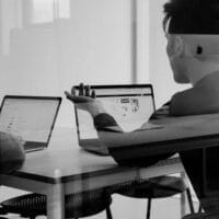 Twee mensen zitten aan een tafel met laptops en bespreken informatie die op hun scherm wordt weergegeven in een moderne kantooromgeving.