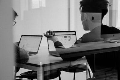 Twee mensen zitten aan een tafel met laptops en bespreken informatie die op hun scherm wordt weergegeven in een moderne kantooromgeving.